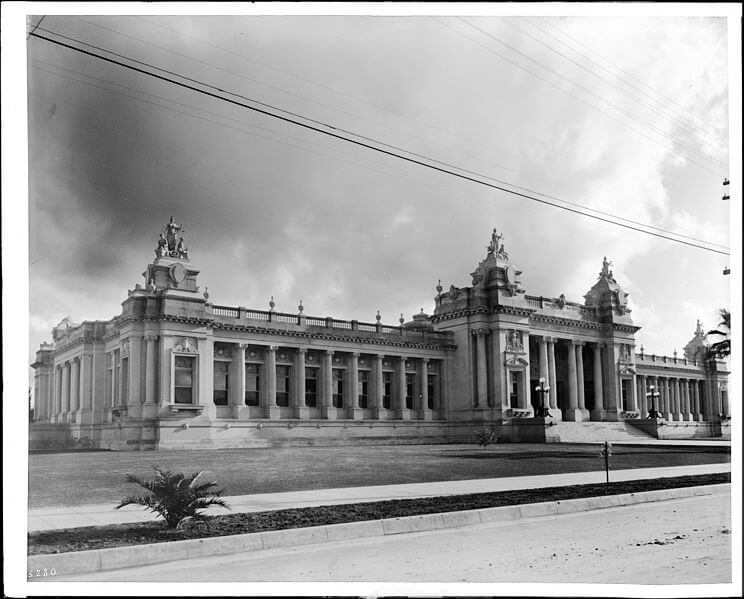 A black and white photograph of a grand neoclassical-style building with large columns, ornate sculptures, and decorative elements along the roofline. The symmetrical structure features an expansive façade with rows of tall windows and a well-manicured lawn in the foreground. Overhead power lines stretch across the sky, and dramatic clouds add depth to the scene, suggesting a historical or civic landmark