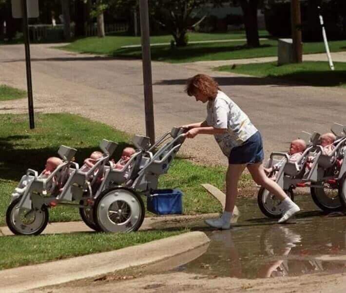 A woman in shorts and sneakers pushes a massive multi-seat stroller carrying several babies across a flooded curb. The front wheels of the stroller are lifted as she struggles to navigate the puddle, while the babies sit strapped in, some looking forward and others gazing around.