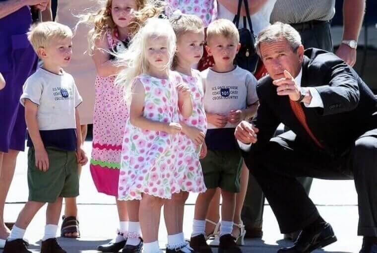 A group of young children, dressed in matching outfits, stand together as former U.S. President George W. Bush crouches beside them, pointing at something in the distance. The children, some in floral dresses and others in khaki shorts with matching shirts, appear curious and slightly puzzled while adults stand in the background.