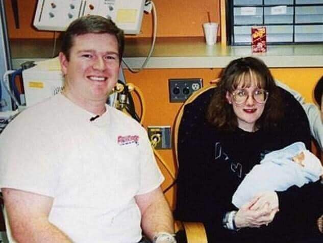 A man in a white t-shirt smiles while sitting next to a woman with glasses, who is holding a newborn baby wrapped in a blue blanket. The setting appears to be a hospital room, with medical equipment, power outlets, and a cup on a shelf in the background.