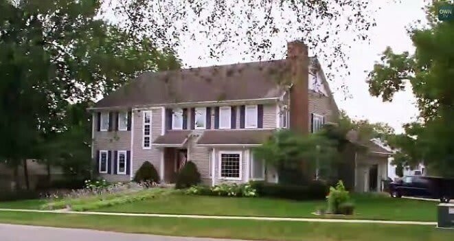 A large two-story suburban home with beige siding, white trim, and a brick chimney sits on a well-manicured lawn with neatly trimmed bushes and flower beds. The house has multiple windows with dark shutters, a small front porch, and a driveway leading to a garage on the right side.