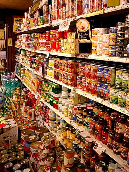 A grocery store aisle filled with shelves stacked high with canned goods, jars, and packaged food items. Various brands and labels of vegetables, beans, sauces, and meats are neatly arranged, with additional cans stacked on the floor.