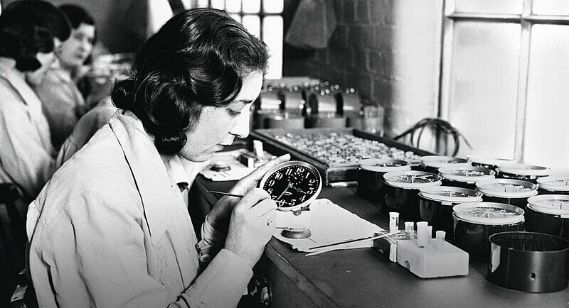 Black and white photo of a woman carefully painting or inspecting the dial of a clock at a workbench. Several other workers are in the background, and multiple clock dials are arranged on the table. The setting suggests an early 20th-century factory or watchmaking workshop."