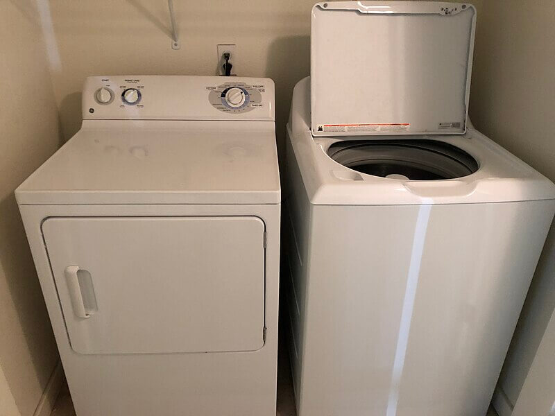 A white top-loading washing machine with its lid open sits next to a white front-loading dryer in a small laundry area. The machines have simple dials for controls, and the space is clean with a power outlet visible on the wall behind them.