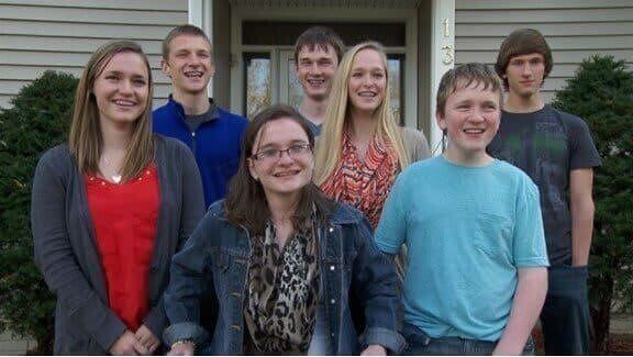 A group of seven smiling teenagers stand together in front of a suburban home with beige siding and green bushes. They are dressed casually, with one girl in a wheelchair at the center, all looking happy and relaxed.