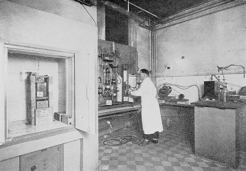 A black and white photograph of a scientist in a lab coat working with complex equipment in a laboratory. The room contains various scientific instruments, pipes, and electrical wiring. A lead-shielded cabinet on the left holds labeled containers, possibly containing radioactive materials, suggesting a mid-20th-century radiochemistry or nuclear research facility