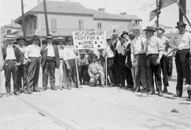 A black and white historical photograph of a group of men standing on a street, holding a sign that reads 'HELP US WIN OUR FIGHT FOR A LIVING WAGE.' Many of them wear hats and suspenders, and some hold American flags. The scene appears to be from an early 20th-century labor protest or workers' strike, advocating for fair wages and better working conditions