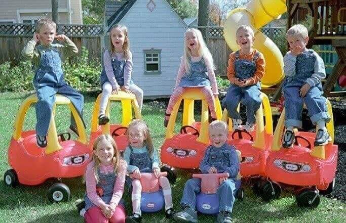 Eight young children dressed in overalls and colorful shirts play in a backyard, sitting on or inside red and yellow toy cars. Some children laugh and pose excitedly, while others sit on small ride-on toys, with a playhouse and slide visible in the background.