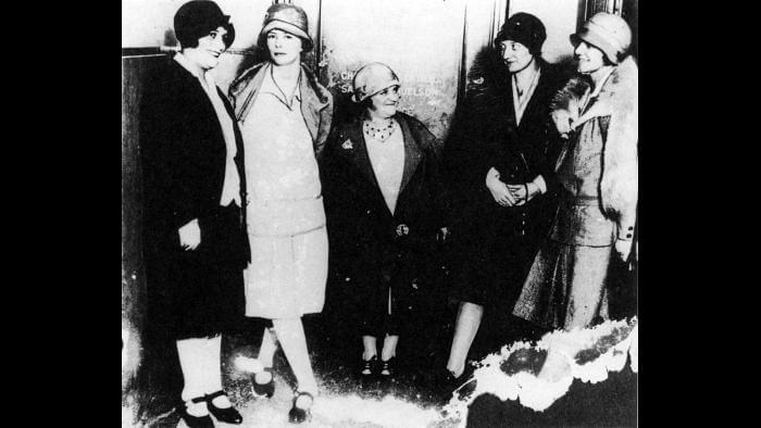 A black and white photograph of five women from the early 20th century, dressed in 1920s-style clothing with cloche hats and knee-length coats. They stand together in conversation, with one noticeably shorter woman in the center. The image appears aged, with visible damage along the edges.