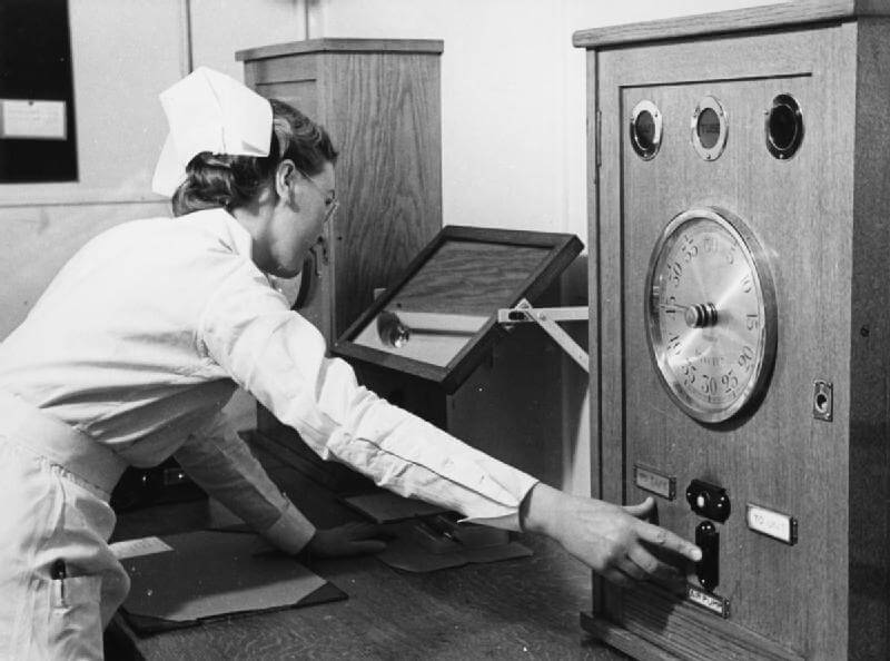 A black and white photograph of a nurse in a traditional uniform and cap operating a large medical or scientific device with a clock-like dial and control switches. She is reaching out to adjust a setting, with paperwork and other equipment visible on the desk beside her. The image suggests a mid-20th-century medical or radiology setting.