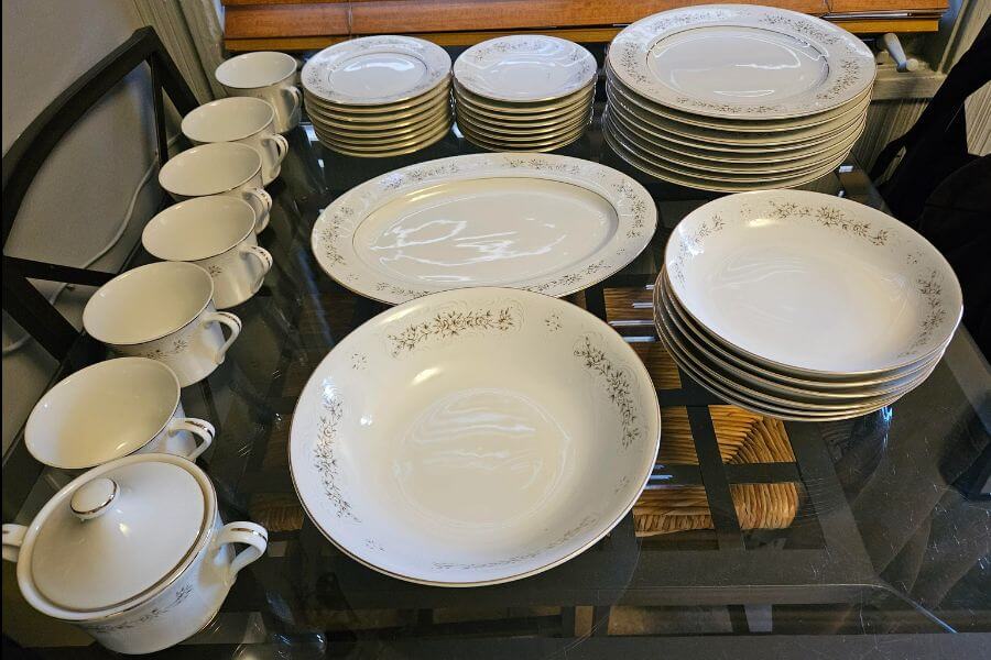 The photograph shows an elegant vintage china set neatly arranged on a glass table. Delicate floral patterns and gold trim adorn the white porcelain, exuding timeless sophistication. Stacks of plates, bowls, and matching cups suggest this set is ready for a formal gathering. A small lidded sugar bowl sits nearby, completing the classic dinnerware ensemble with refined charm.





