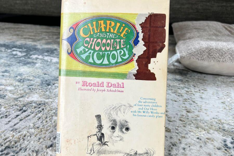 The photograph shows a well-loved copy of Charlie and the Chocolate Factory by Roald Dahl resting on a textured rug. Its cover features the iconic title in a colorful, whimsical font alongside an illustration of Willy Wonka and a melting chocolate bar. The book’s edges are slightly worn, suggesting it has been cherished and read many times. With its classic storytelling and imaginative world, this novel remains a timeless favorite for readers of all ages.