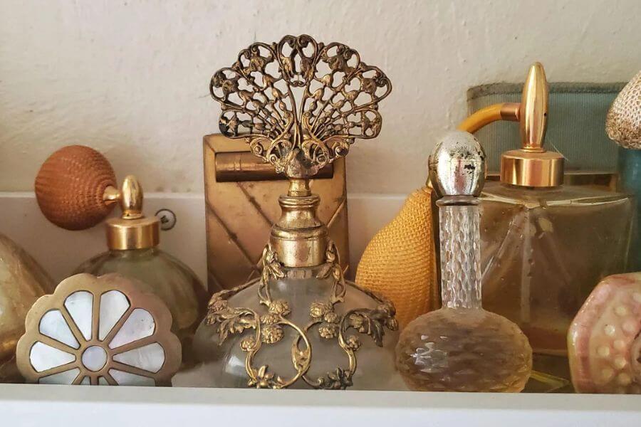 The photograph shows a delicate collection of vintage perfume bottles elegantly displayed on a shelf. Intricate glasswork, ornate gold embellishments, and filigree details make each bottle a small work of art. Some feature atomizer bulbs in rich amber hues, while others have decorative stoppers gleaming with an antique patina. With their timeless beauty, these bottles evoke a sense of old-world glamour and sophistication.