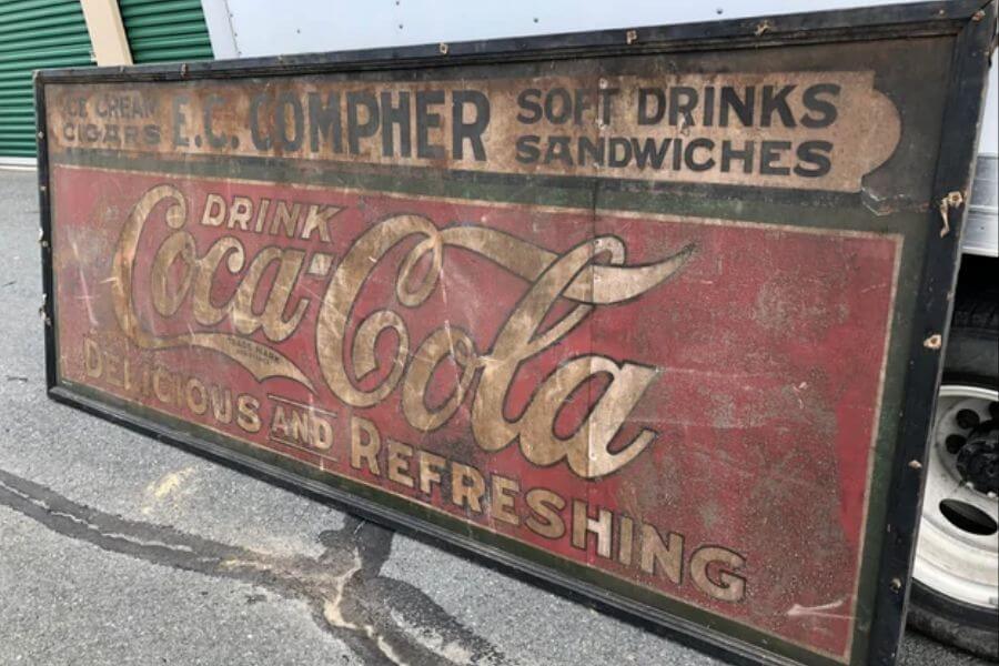 The photograph shows a large, weathered Coca-Cola advertisement sign leaning against a truck. Faded red and gold lettering spell out “Drink Coca-Cola” alongside the slogan “Delicious and Refreshing.” The sign also features an old business name, "E.C. Compher," advertising soft drinks, cigars, and sandwiches. With its chipped paint and rusted edges, this relic tells a story of soda fountains, roadside diners, and a bygone era of classic Americana.