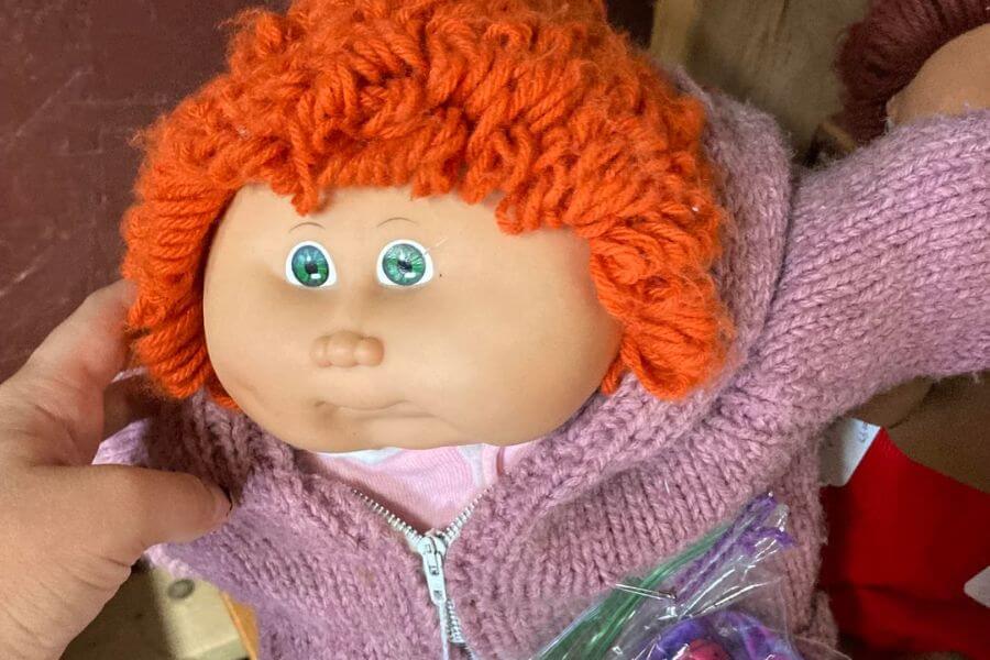 The photograph shows a classic Cabbage Patch Kid doll with bright green eyes and a signature round face. Her curly orange yarn hair is topped with a tiny purple ribbon, adding a touch of charm. Dressed in a cozy pink knitted sweater, she exudes vintage nostalgia and childhood comfort. A small plastic bag of extra doll clothes rests in her lap, ready for a wardrobe change at any moment.