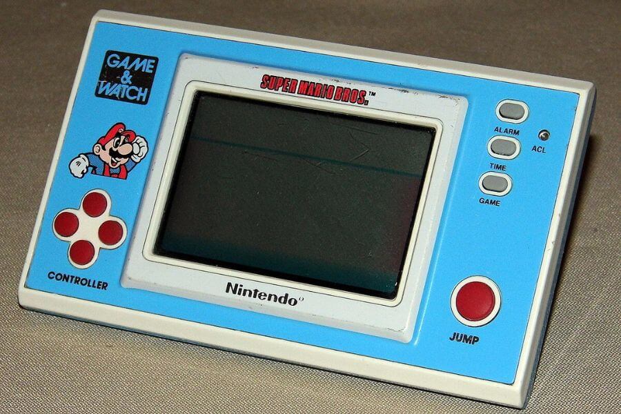 The photograph shows a vintage Nintendo Game & Watch handheld featuring Super Mario Bros. in its classic 1980s design. The compact device has a blue and white color scheme, with a simple LCD screen and bold red buttons. A cheerful Mario graphic in the corner adds charm, reminding players of the golden age of handheld gaming. 