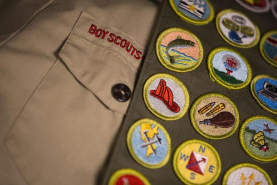 The photograph shows a Boy Scouts uniform neatly laid out, featuring a khaki shirt with red embroidered lettering. A green sash draped over the shirt is adorned with a colorful collection of merit badges, each representing a different skill or achievement. From fire safety to navigation, these small circular patches tell a story of dedication and adventure. 