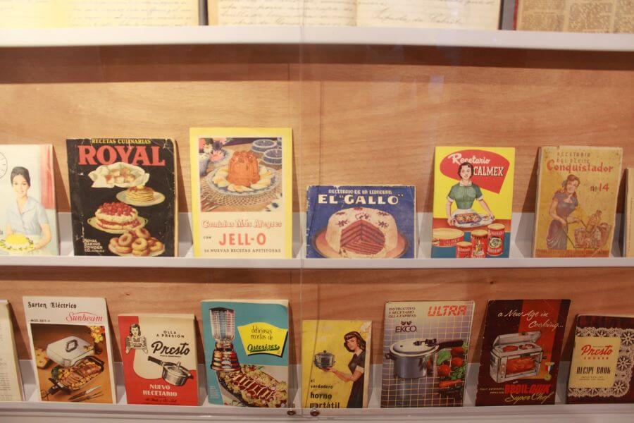 The photograph shows a carefully arranged display of vintage cookbooks and appliance manuals, neatly placed behind glass. Their colorful covers feature nostalgic illustrations of homemakers, classic kitchen appliances, and beautifully plated dishes from mid-century kitchens. Titles like Royal Baking Powder, Jell-O Recipes, and Presto Pressure Cookers hint at a time when convenience cooking was on the rise.