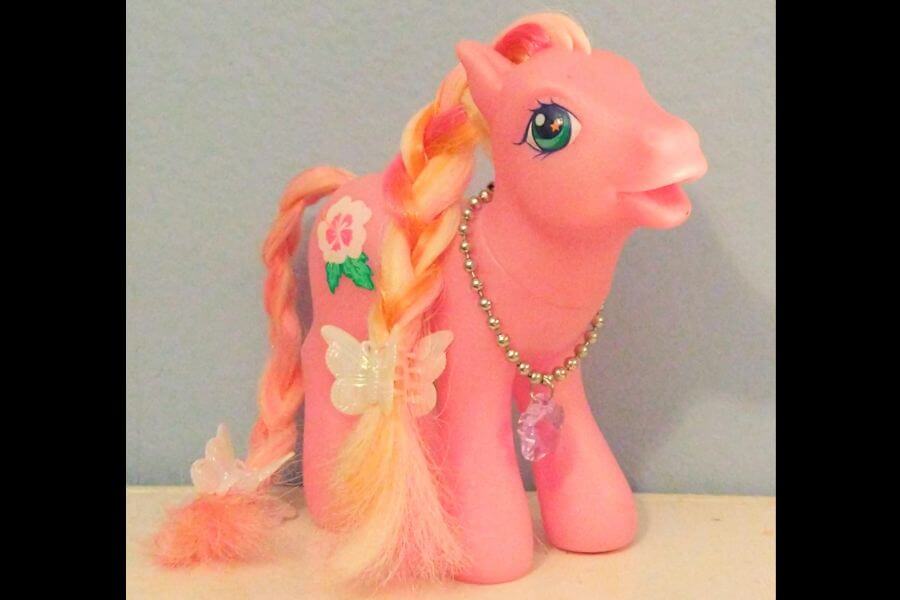 The photograph shows a vintage My Little Pony figure in a soft pastel pink hue. Her bright green eyes sparkle, framed by long lashes and a joyful expression. A silky mane of pink and yellow is braided neatly, adorned with translucent butterfly clips. Around her neck, a tiny beaded necklace with a purple crystal pendant adds a touch of magical charm.