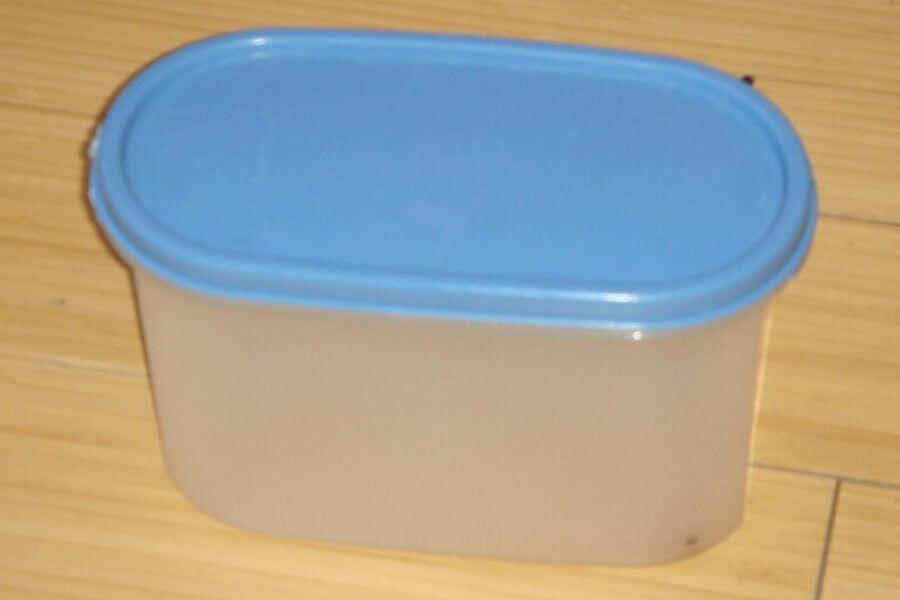 The photograph shows a vintage plastic storage container with a light blue lid resting on a wooden floor. Its semi-transparent body suggests it was once a kitchen essential, likely used for storing dry goods or leftovers. The snug-fitting lid and classic design hint at a well-loved piece of Tupperware history. 