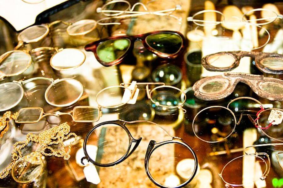 The photograph shows a collection of vintage eyeglasses displayed on a reflective glass surface. A mix of round wire frames, tinted lenses, and antique pince-nez styles showcase different eras of optical fashion. Among them, a worn pair of leather-bound spectacles stands out, hinting at a time before modern eyewear.