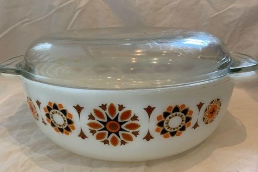 The photograph shows a vintage Pyrex casserole dish, featuring a bold mid-century floral pattern. Warm tones of orange, brown, and white create a striking geometric design on the milky glass surface. A clear glass lid rests on top, showcasing the classic durability and functionality of this kitchen staple. 