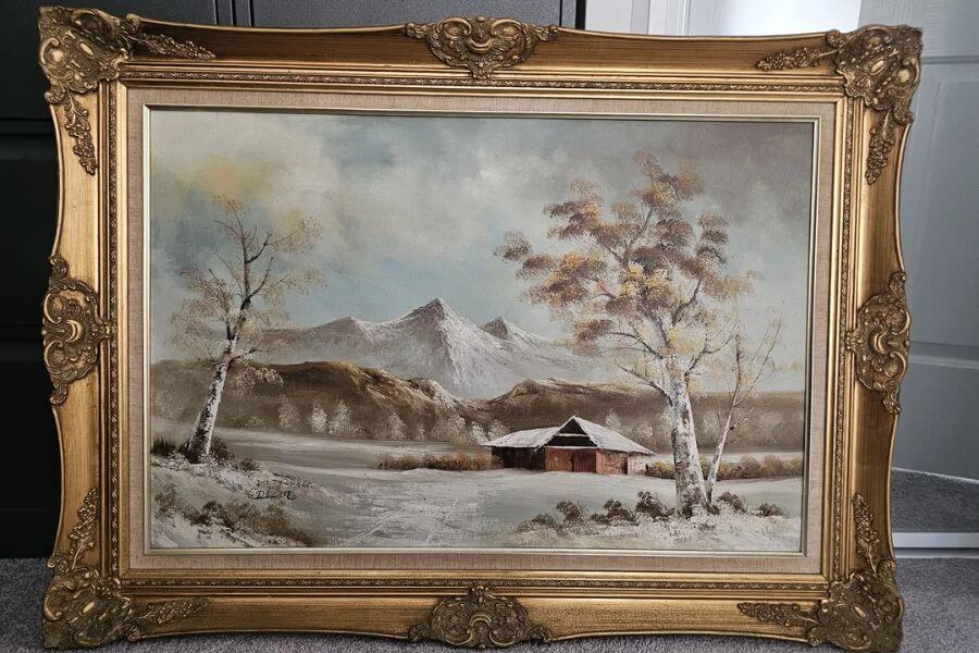 The photograph shows a framed landscape painting, capturing a serene winter scene in a rustic countryside. The painting features snow-dusted trees, a cozy wooden cabin, and towering mountains, set against a soft sky with muted blues and grays. The ornate gold frame, with its elaborate carvings and baroque-style flourishes, adds a touch of grandeur to the artwork. 