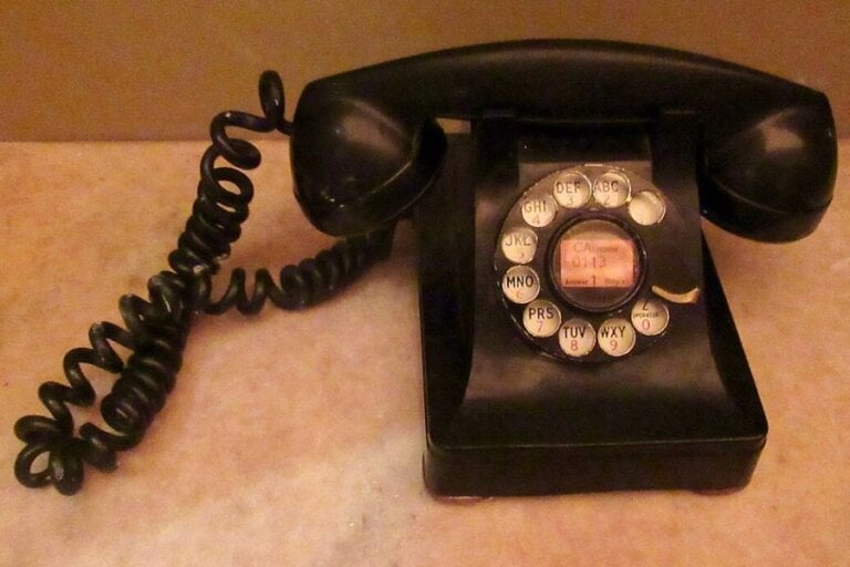 The photograph shows a vintage black rotary telephone resting on a marble surface. Its coiled cord stretches out, slightly tangled, hinting at years of use. The numbered dial, complete with a worn-out center label, recalls a time when dialing required patience. With its sturdy handset and timeless design, this phone once connected voices across distances before touchscreens took over.