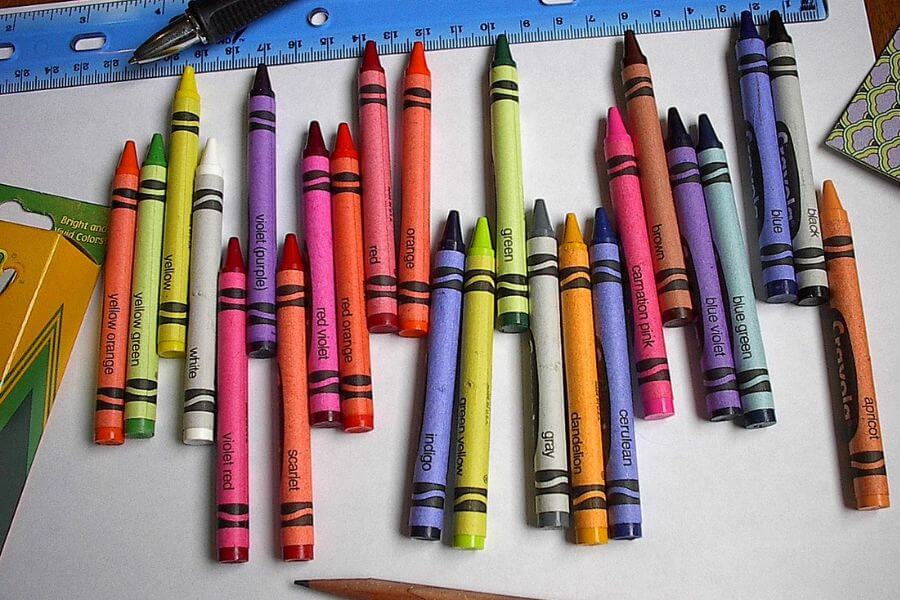 The photograph shows a scattered collection of colorful Crayola crayons sprawled across a white sheet of paper. Their labeled wrappers proudly display shades like "cerulean," "scarlet," and "blue violet." A worn-down wooden pencil rests nearby, looking slightly neglected next to the vibrant wax army. With a ruler and a pen in the background, this scene feels like the calm before a creative storm.