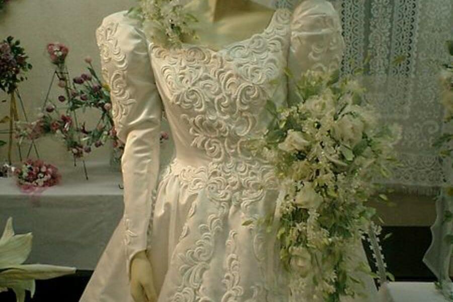 The photograph shows an elegant mannequin dressed in a vintage-style wedding gown with intricate lace embroidery. Puffy sleeves and a voluminous skirt add a dramatic, fairy-tale quality to the ensemble. A cascading bouquet of white flowers drapes gracefully over the mannequin’s arm, enhancing the bridal aesthetic. Behind it, lace decorations and floral arrangements complete the romantic, old-fashioned ambiance.