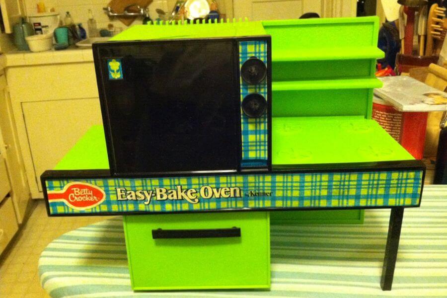 The photograph shows a vintage Betty Crocker Easy-Bake Oven by Kenner, standing proudly in its bold lime-green glory. Blue plaid accents give it a retro charm, making it a nostalgic gem from the past. The black oven door, two control knobs, and side counter with built-in shelving complete its miniature kitchen design. This beloved toy once let kids whip up tiny treats using nothing but the power of a light bulb.