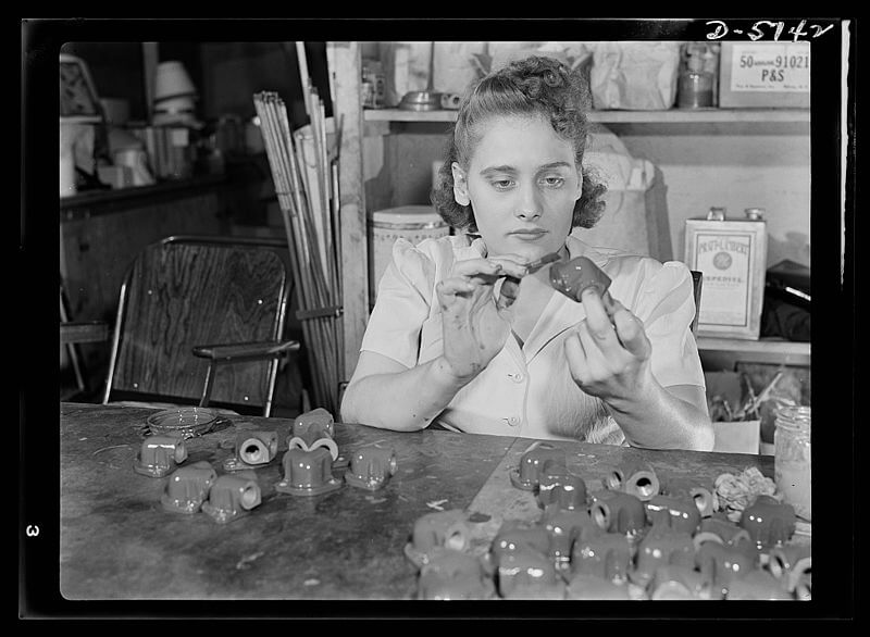 Black and white photo of a woman in a workshop carefully painting or inspecting a small object. Several similar objects are spread out on the table in front of her. Shelves in the background hold tools, containers, and supplies, suggesting an industrial or manufacturing setting