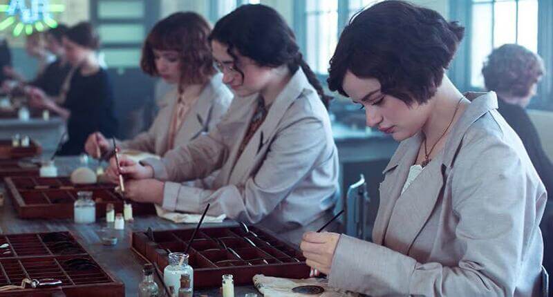A colorized or reenacted historical image of young women in a factory setting, dressed in early 20th-century attire, painting watch dials with fine brushes. They are seated at long worktables with small bottles of paint and organized compartments for materials. The scene closely resembles the working conditions of the 'Radium Girls,' who unknowingly handled radioactive paint.
