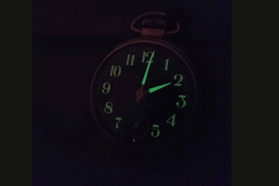 A photograph of a vintage pocket watch glowing in the dark, displaying luminescent numbers and hands. The greenish glow suggests the use of radium-based paint, historically used in early 20th-century timepieces for visibility in low light. The watch face is partially obscured by darkness, with the time appearing to be around 11:55.