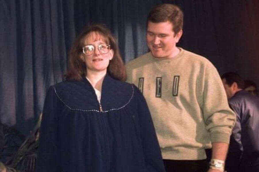 A  woman wearing glasses and a dark blue oversized gown walks forward with a neutral expression, while a man in a beige "NED" sweatshirt smiles behind her. He wears a hospital bracelet on his wrist, and the background features a dark curtain with other people present.