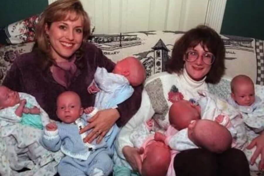 Two women sit on a patterned couch, each holding multiple newborn babies dressed in pastel-colored outfits. One woman, wearing a dark jacket and scarf, smiles brightly, while the other, in glasses and a sweater, holds several infants with a more subdued expression.