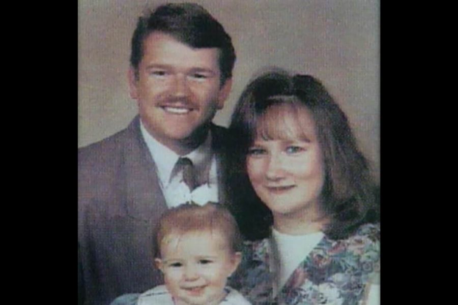 A vintage-style family portrait featuring a father, mother, and baby. The father wears a suit and tie with a mustache, the mother has shoulder-length hair with bangs, and the baby, dressed in a light-colored outfit with a bow, sits in front, smiling.