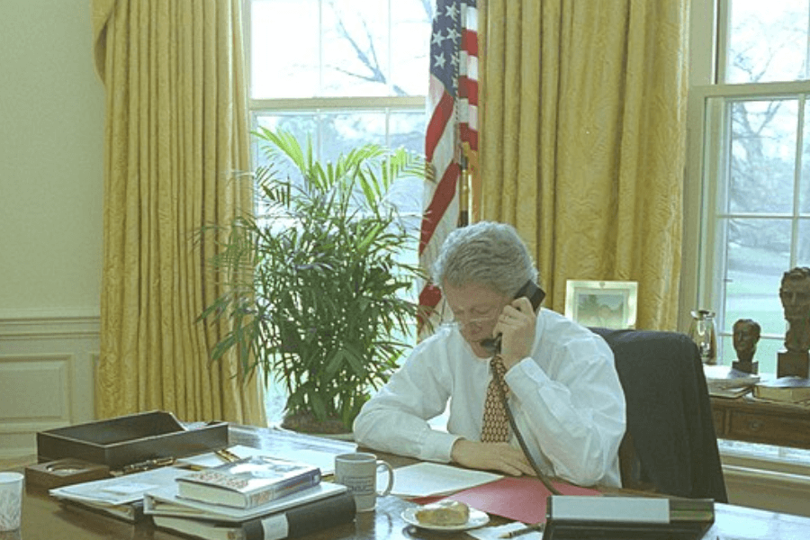 Former U.S. President Bill Clinton sits at the Resolute Desk in the Oval Office, speaking on the phone while reviewing documents. 