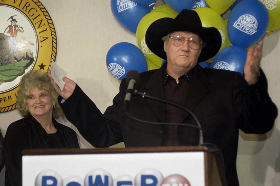 A man in a black cowboy hat and dark suit stands at a podium with "Powerball" written on it, holding up a piece of paper while gesturing with both hands. He has a neutral expression and wears glasses. Behind him, a smiling woman with curly blonde hair stands next to a West Virginia state seal, with blue and yellow balloons displaying the "West Virginia Lottery" logo in the background.