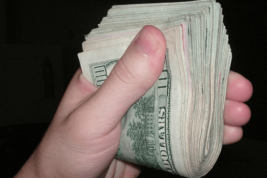 A close-up of a hand tightly gripping a thick stack of U.S. $100 bills. The edges of the bills appear slightly worn, and the top bill prominently displays "100 DOLLARS." The background is dark and out of focus.