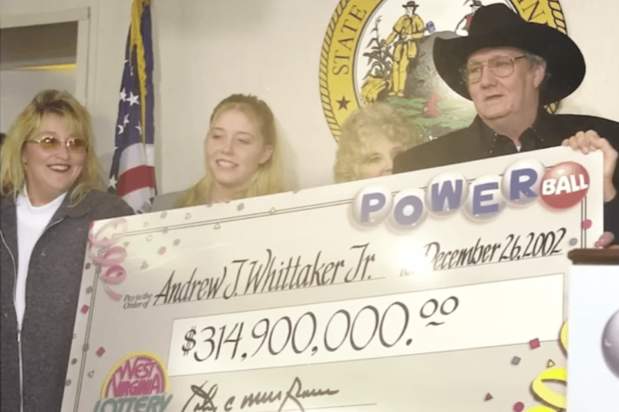 A group of people stands together holding an oversized Powerball lottery check for $314.9 million, dated December 26, 2002. The check is made out to Andrew J. Whittaker Jr., with the "West Virginia Lottery" logo visible. A man in a black cowboy hat and glasses, appearing to be the winner, stands at the podium while smiling women stand beside him. An American flag and the West Virginia state seal are displayed in the background.