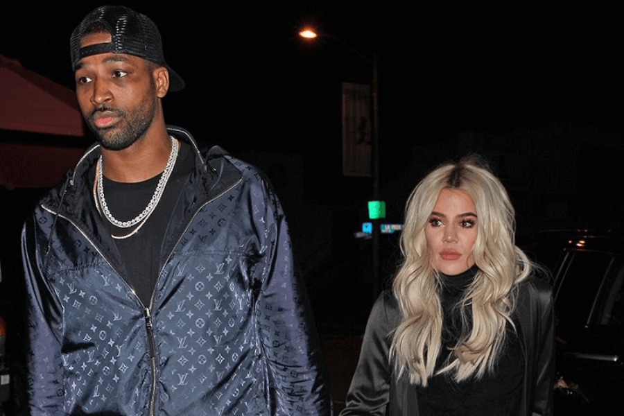 A man wearing a black Louis Vuitton jacket, a backward cap, and a silver chain walks beside a woman with long, wavy blonde hair. The woman, dressed in a black satin outfit, has a neutral expression with a slightly serious gaze. The nighttime setting includes streetlights and a dark background with some illuminated signs.