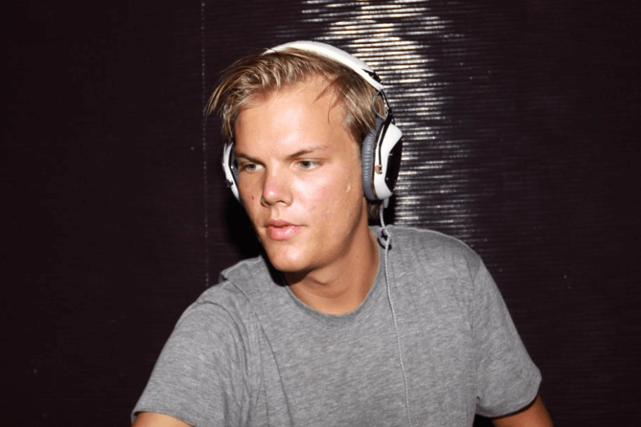 A young male DJ with short blonde hair and a focused expression wears white over-ear headphones while performing. He is dressed in a casual gray t-shirt, with dim lighting casting a glow on his face. The dark, textured background suggests a club or music venue setting.