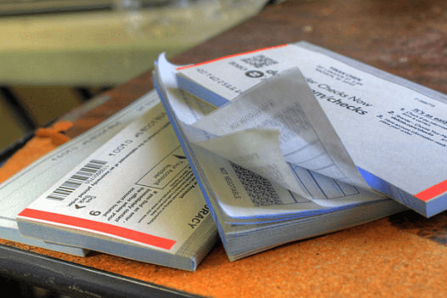 A close-up of several checkbooks resting on a worn wooden surface, with one open showing carbon copies of previously written checks. The checkbooks have security features, barcodes, and printed instructions on the covers.