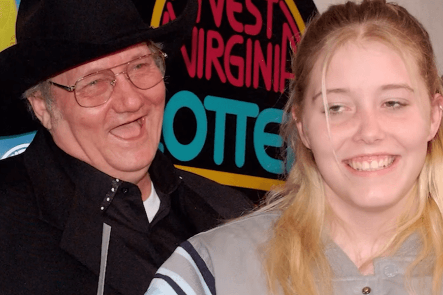 A man in a black cowboy hat and glasses laughs joyfully while standing next to a young blonde woman who is smiling. Behind them, a colorful "West Virginia Lottery" sign is visible, suggesting a celebratory event. The man appears to be dressed in a formal black outfit, while the woman wears a casual jacket.