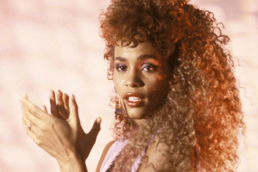 A glamorous female singer with voluminous curly hair and bold 80s-style makeup claps her hands while looking towards the camera. She wears statement earrings and a sparkling outfit, with warm, soft lighting enhancing the retro aesthetic. Her confident expression and dynamic pose capture the energy of a performance.