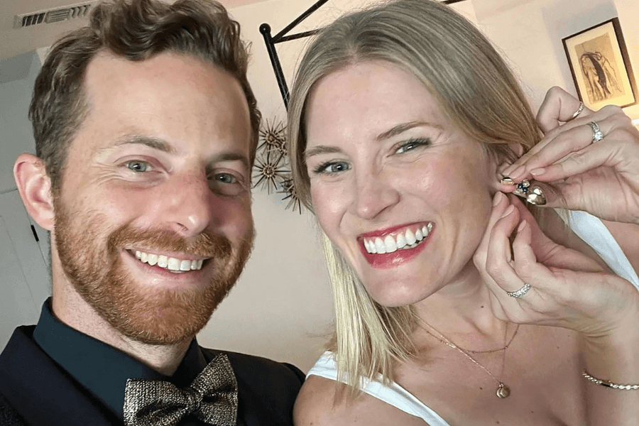 A smiling man with short, curly red hair and a beard wears a black dress shirt with a gold bow tie, posing for a selfie. Beside him, a blonde woman with bright red lipstick and a white top grins while playfully holding up a colorful earring to her ear. The background features modern home decor, including framed artwork and decorative wall accents.