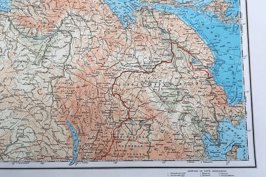 A detailed, vintage topographic map displaying a region with various elevations, rivers, and marked routes. The map includes Russian text, with red boundary lines and contour shading indicating terrain features. A portion of a blue coastline is visible on the right side.