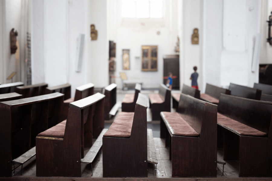 A quiet, empty church interior with rows of dark wooden pews lined with faded red cushions. The high white walls and tall windows allow natural light to stream in, creating a serene atmosphere. In the background, two children can be seen walking near religious artwork and statues.