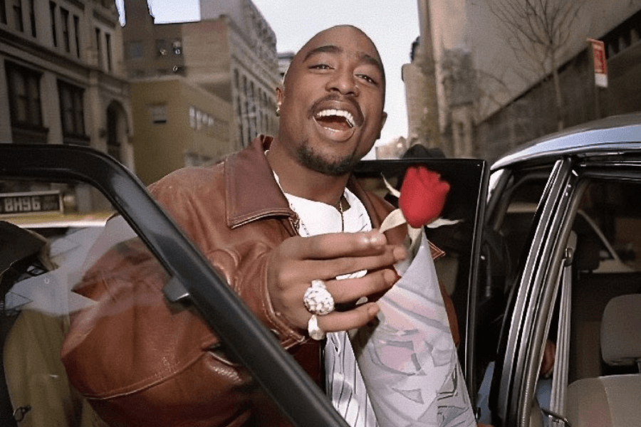 A man with a shaved head and goatee smiles warmly while stepping out of a car, extending a single red rose toward the camera. He wears a brown leather jacket over a white striped shirt, accessorized with large rings and a gold chain. The urban backdrop features tall buildings and city streets, adding a vibrant, candid feel to the moment.